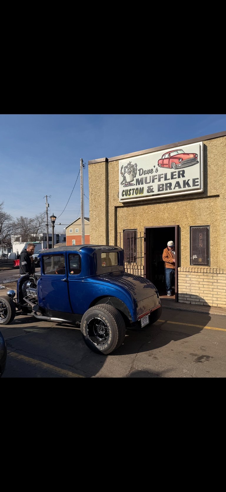 Blue vintage hot rod parked outside Daves Muffler Custom & Brake shop with man standing in doorway