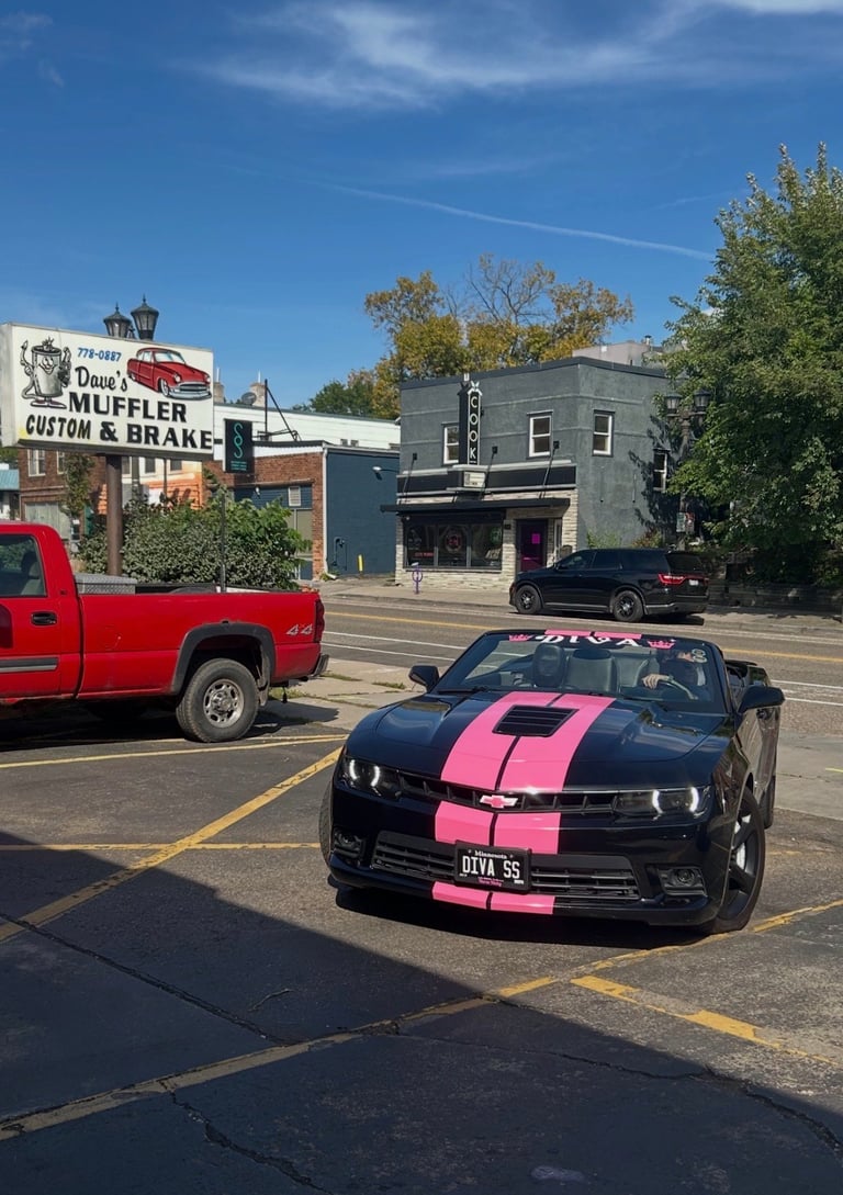 Black Mustang with pink racing stripes parked on city street with vintage diner sign and red truck visible