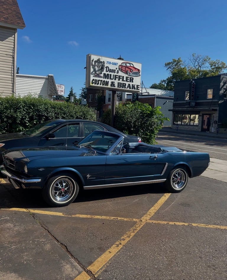 Blue 1960s Ford Mustang convertible parked in front of a custom car shop with sign visible